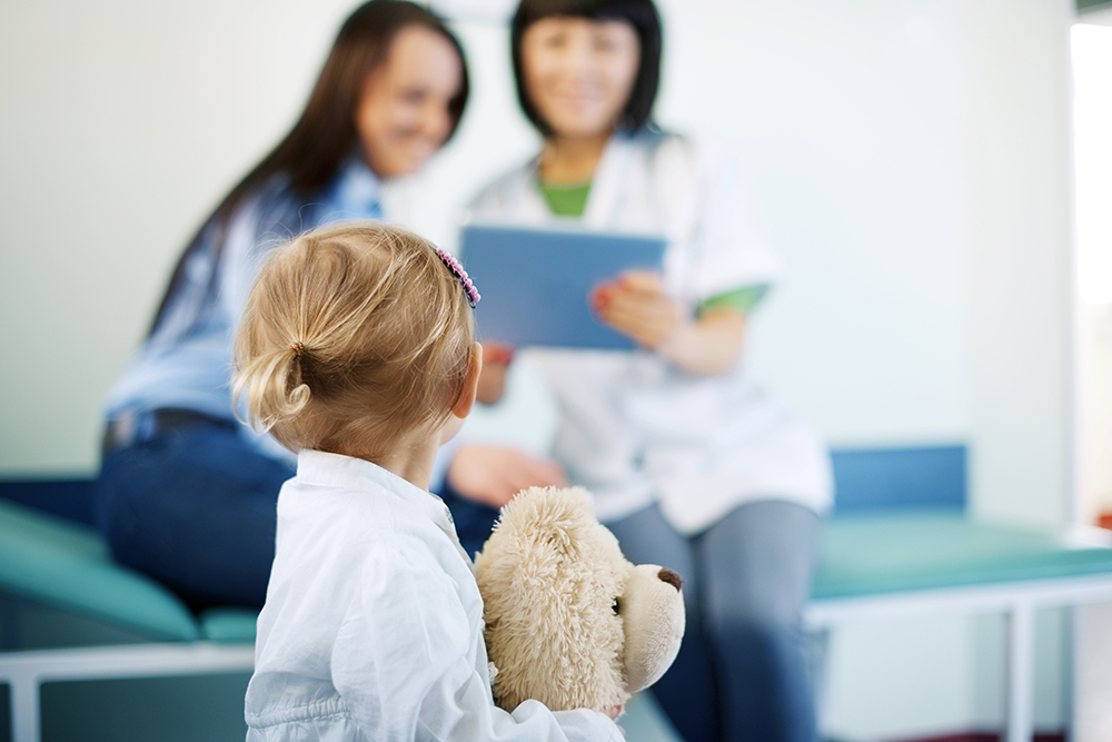 A child on his back holding a teddy bear. In the background is a woman sitting next to a health professional. They are sitting and looking at a clipboard that the health worker is holding.