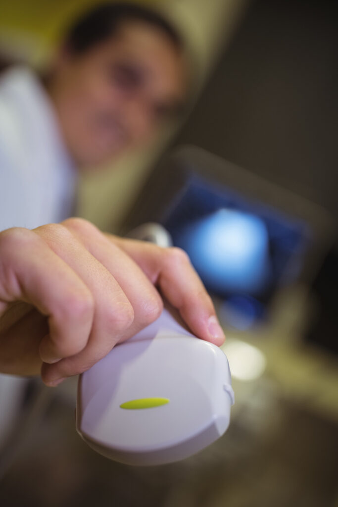 Médico segurando um transdutor de ultrassom. O foco da imagem está em sua mão; o corpo e o computador ao fundo exibindo a imagem de ultrassom estão desfocados.